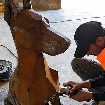 A craftsman using an angle grinder to finish a faceted animal figure built from welded weathering-steel plates in the workshop