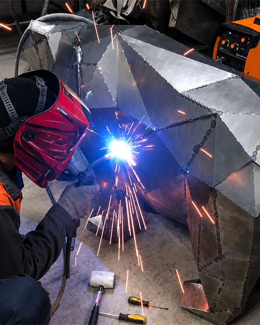 A welder in a red helmet arc-welding the seams of a large faceted steel sculpture, with bright sparks in the workshop