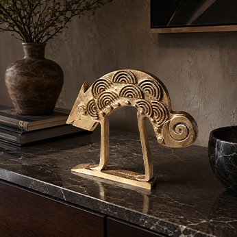 Small handcrafted animal figure in a gold finish with concentric-spiral surface motifs and a curled tail, displayed on a dark marble console in a styled interior.