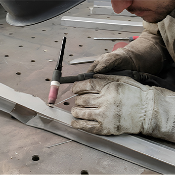 Close-up of a gloved hand TIG-welding a metal seam beside a perforated steel fixture table in the sculpture workshop.